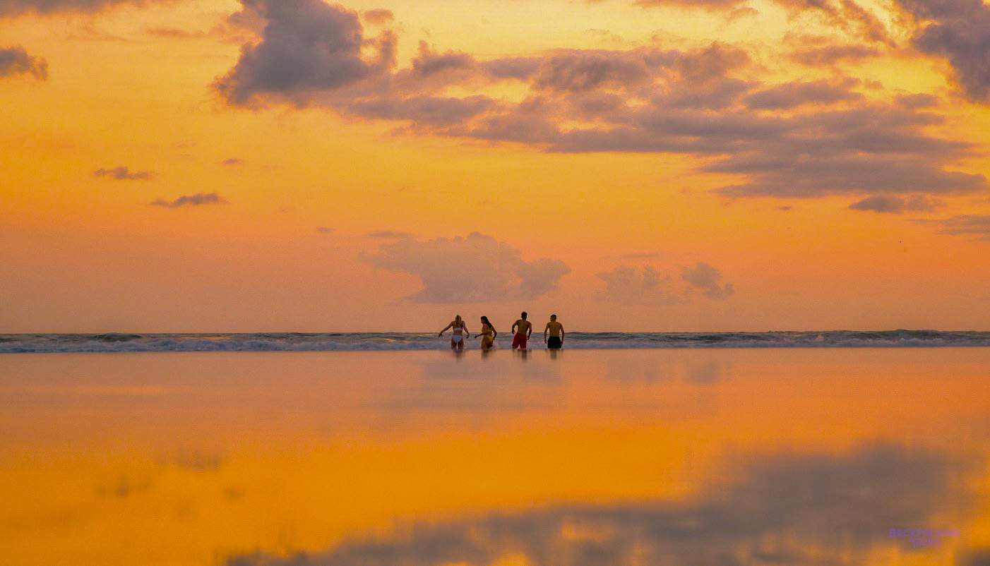 Four people wade in the shallow, mirror-like waters of Gili Trawangan under a vibrant sunset sky.