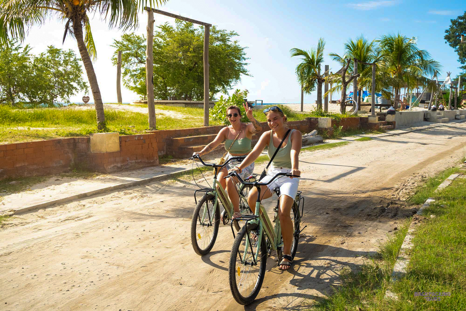 Two happy women cycling along a sandy path on Gili Trawangan with palm trees and a beach.