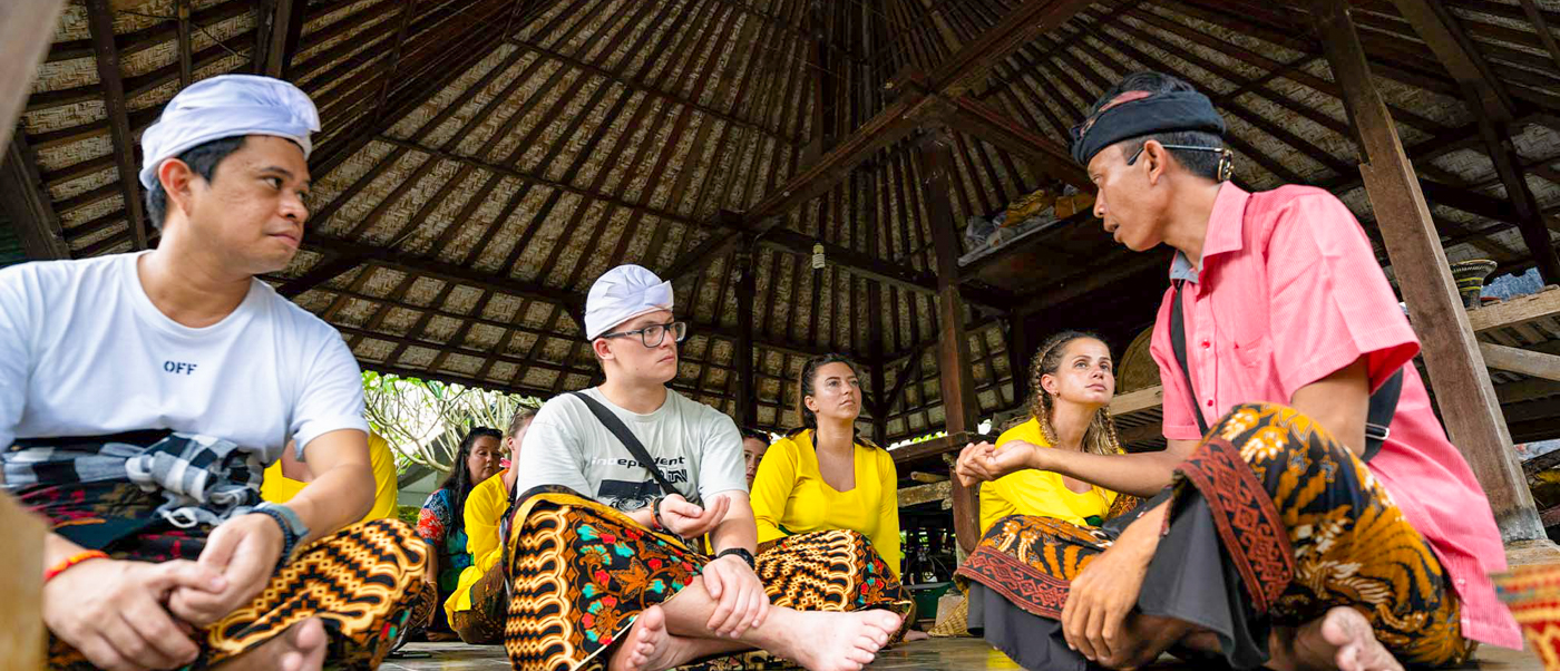 Travelers in Bali respectfully learn from a local guide inside a traditional pavilion.
