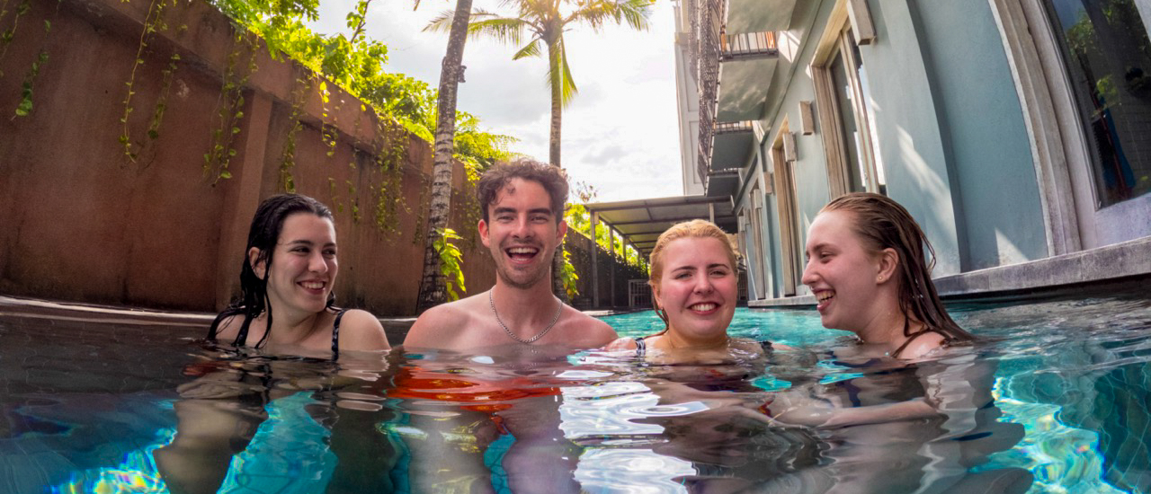 Four smiling friends enjoy a refreshing swim in a sunny hotel pool in Canggu, Bali.