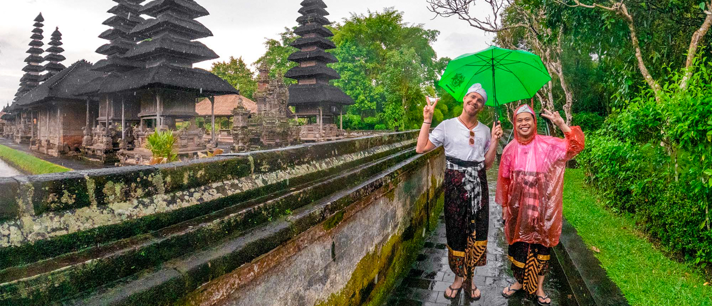 Two smiling people in traditional Balinese sarongs visit an ancient temple in Canggu during rain.
