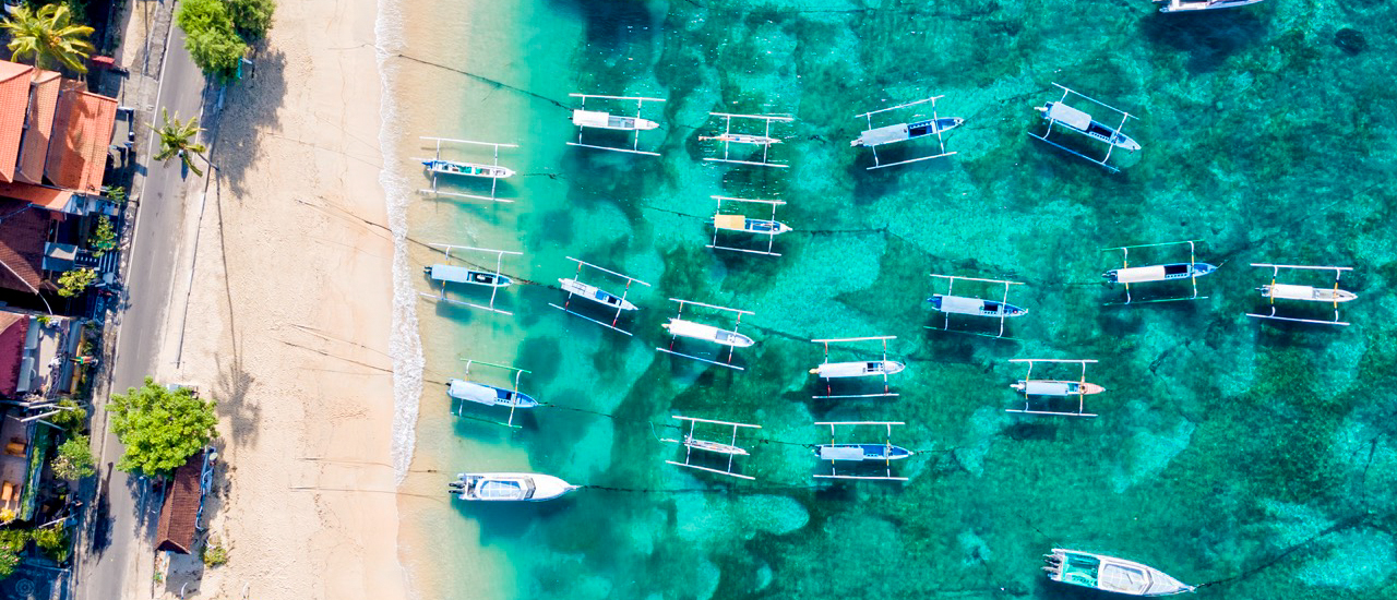 Aerial view of many traditional boats in clear turquoise water next to a sandy beach and coastal buildings.