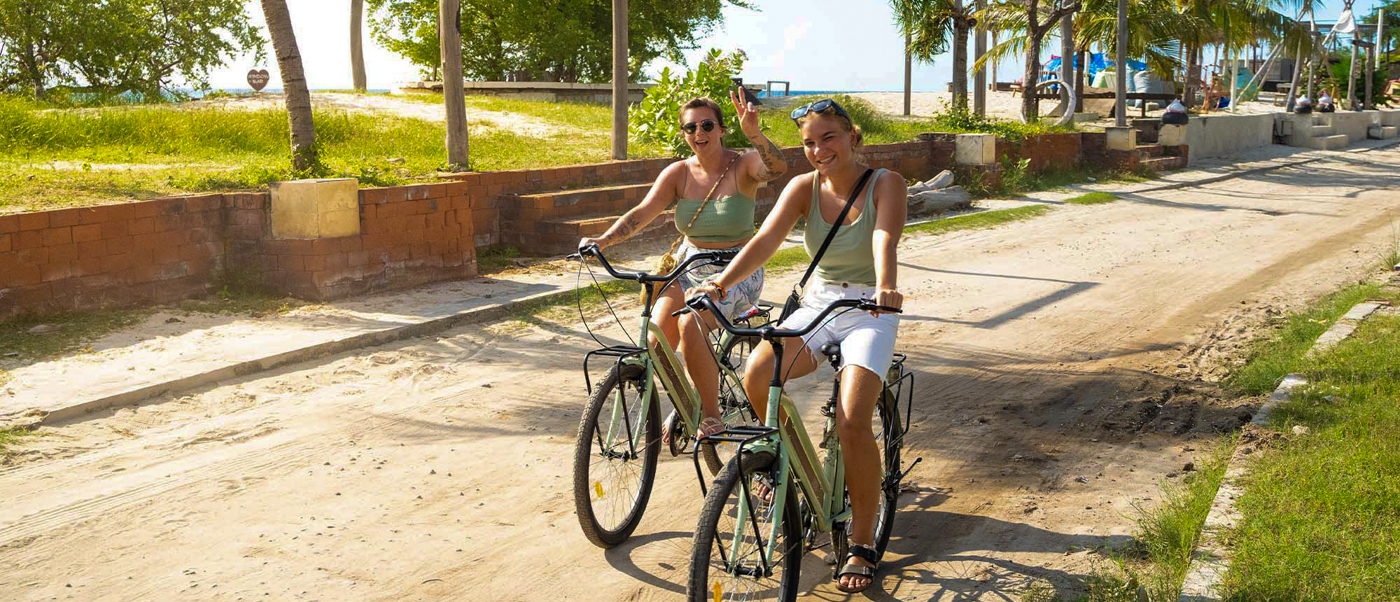 Two smiling women ride bicycles on a sandy path with palm trees on Gili Trawangan.