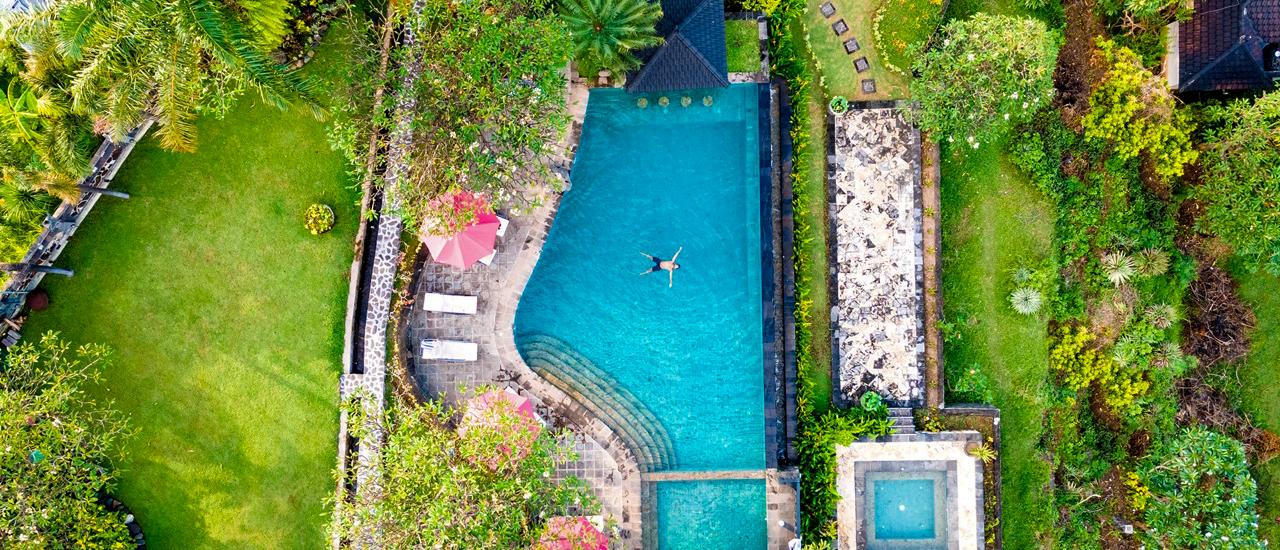 A person floats in a luxurious infinity pool at a villa in Lovina, Bali, amidst lush gardens.