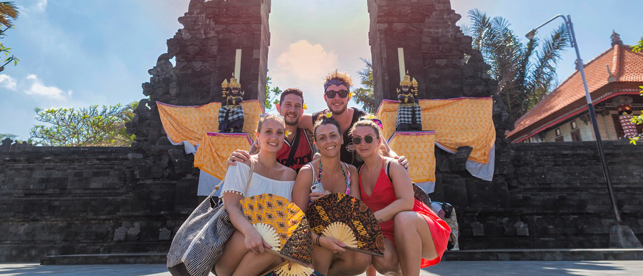 Five happy friends pose with decorative fans in front of a traditional Balinese temple gate.