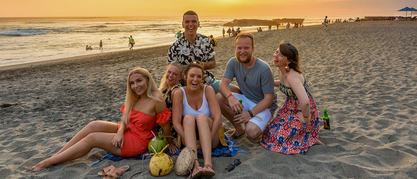 Six happy friends enjoying sunset on a sandy beach in Canggu, Bali, with drinks.
