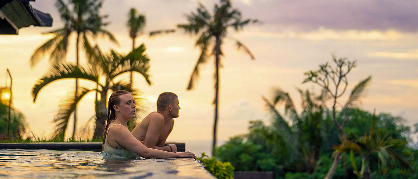A couple relaxes in an infinity pool, enjoying a beautiful tropical sunset over Lovina, Bali.