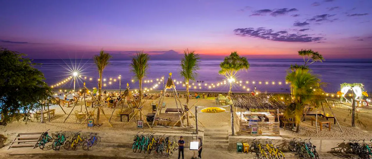 A vibrant beach bar on Gili Trawangan at sunset, with string lights, palm trees, and many bicycles.