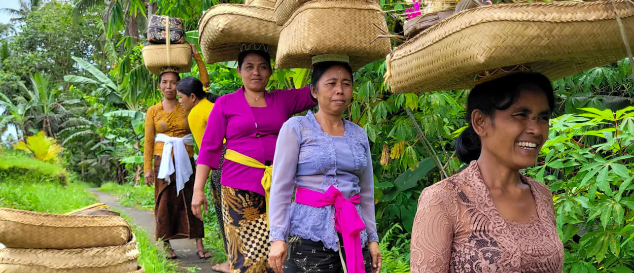 Smiling Balinese women carry traditional woven baskets on their heads through lush jungle in Ubud.