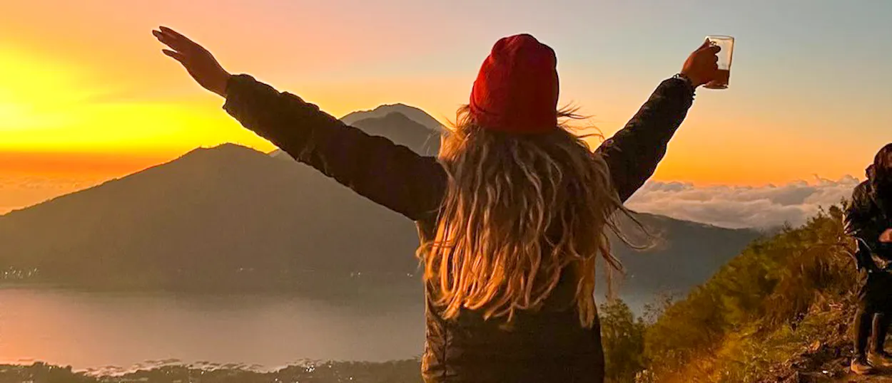A hiker with outstretched arms holds coffee, watching the stunning sunrise over Mount Batur, Bali.