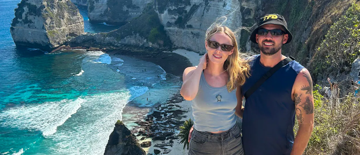 Two travelers smile, enjoying the breathtaking views of Kelingking Beach and its turquoise waters in Nusa Penida.