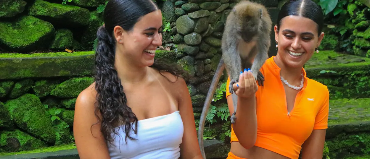 Two happy women interact with a monkey in the lush, moss-covered Ubud Monkey Forest.