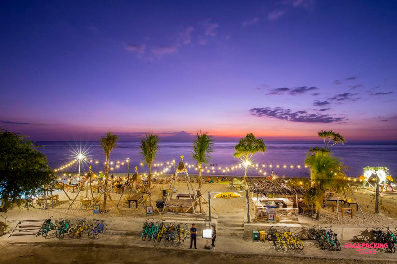 A lively beach bar on Gili Trawangan, illuminated by string lights under a beautiful purple sunset.