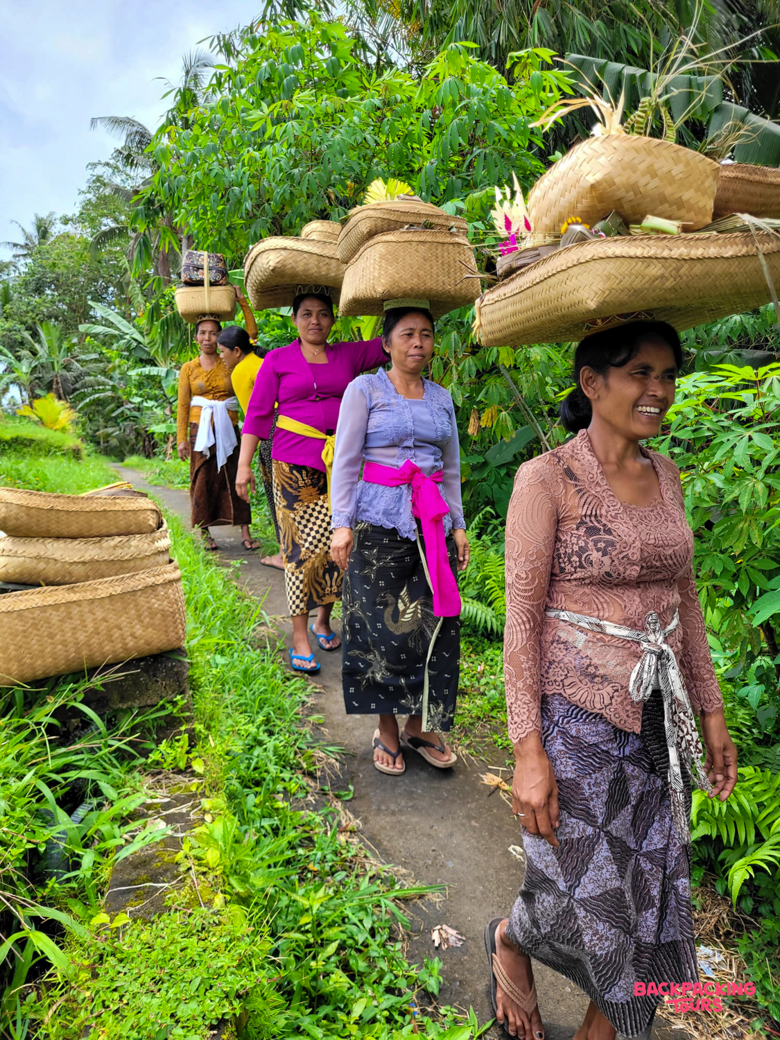 Balinese women in traditional clothing carry woven baskets on their heads along a jungle path in Ubud.