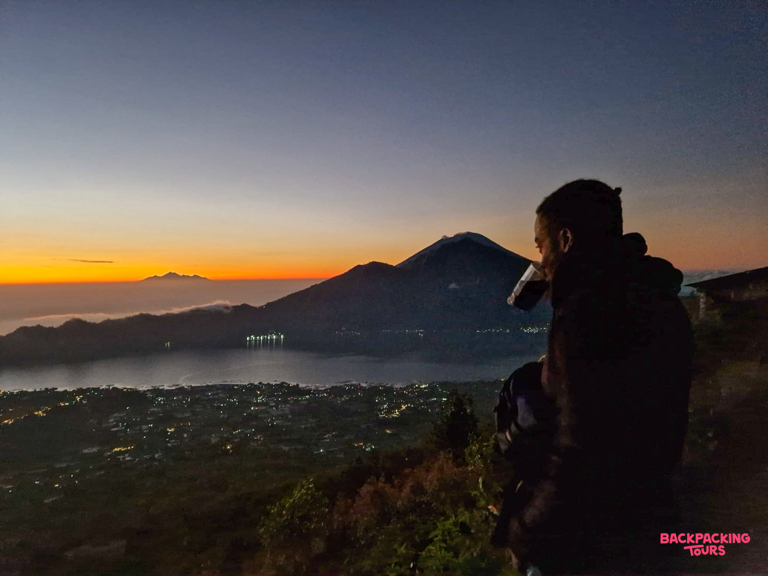 Hiker sips coffee at sunrise, enjoying panoramic views of Bali's volcanic landscape from Mount Batur.