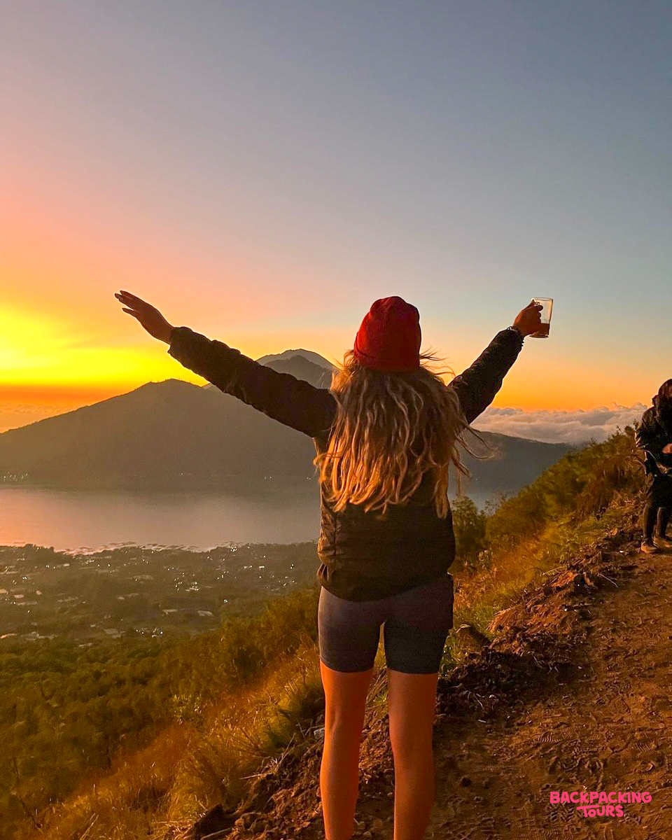 A hiker with outstretched arms sips coffee, enjoying the vibrant sunrise over Mount Batur, Bali.