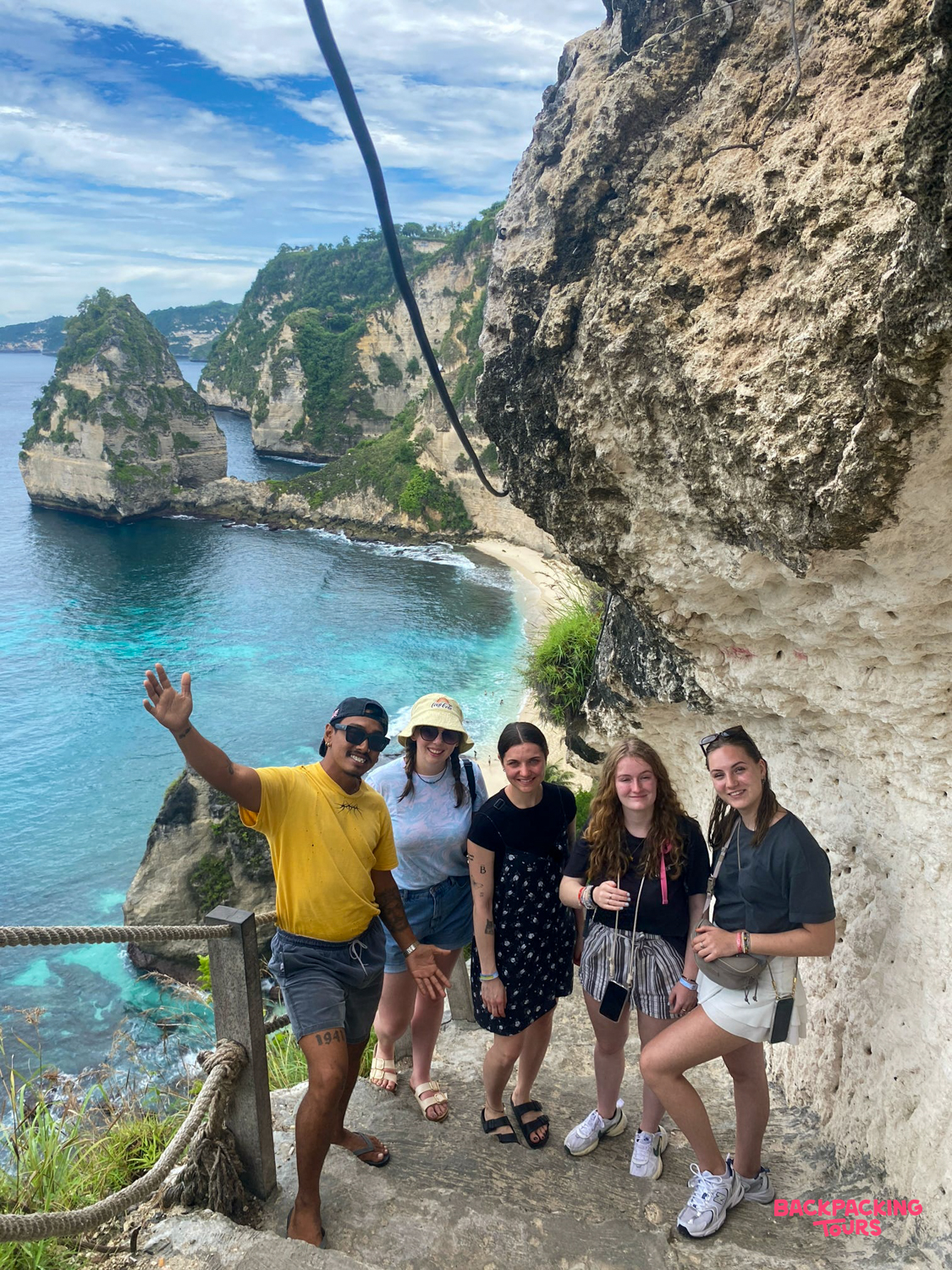 Five happy travelers and a guide pose on a cliffside path overlooking Kelingking Beach, Nusa Penida.