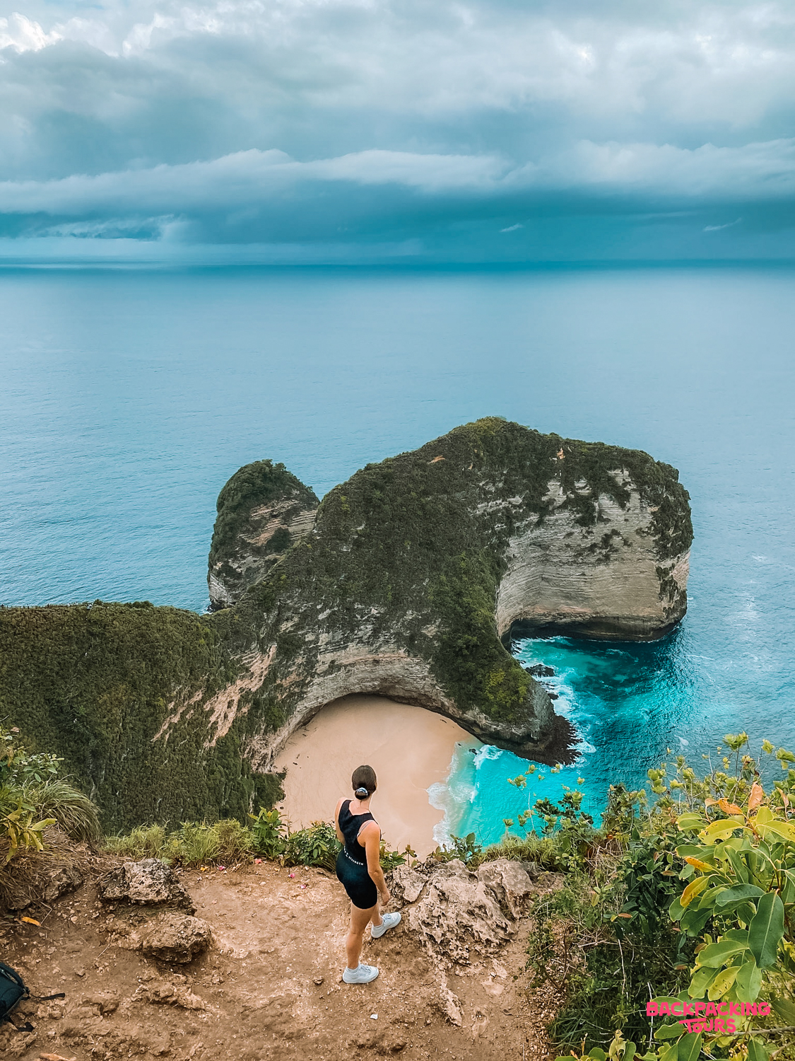 A woman stands on a cliff admiring the iconic T-Rex shaped Kelingking Beach in Nusa Penida.