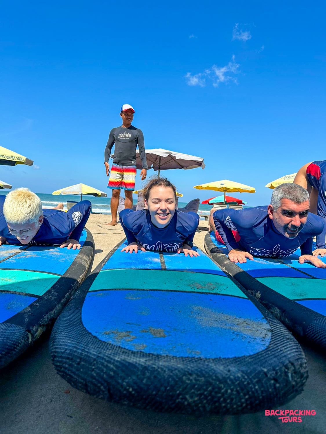 Travelers practice surfing techniques on blue boards on the sand with an instructor in Canggu, Bali.