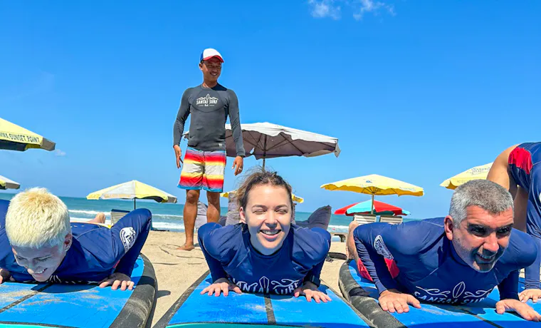 Travelers practice surfing techniques on blue boards on the sand with an instructor in Canggu, Bali.