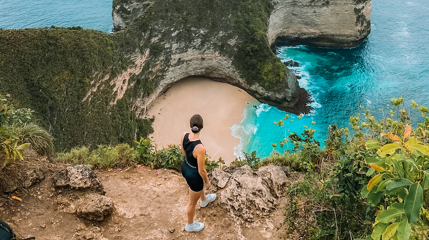 A woman stands on a cliff overlooking the stunning Kelingking Beach with turquoise water in Bali.