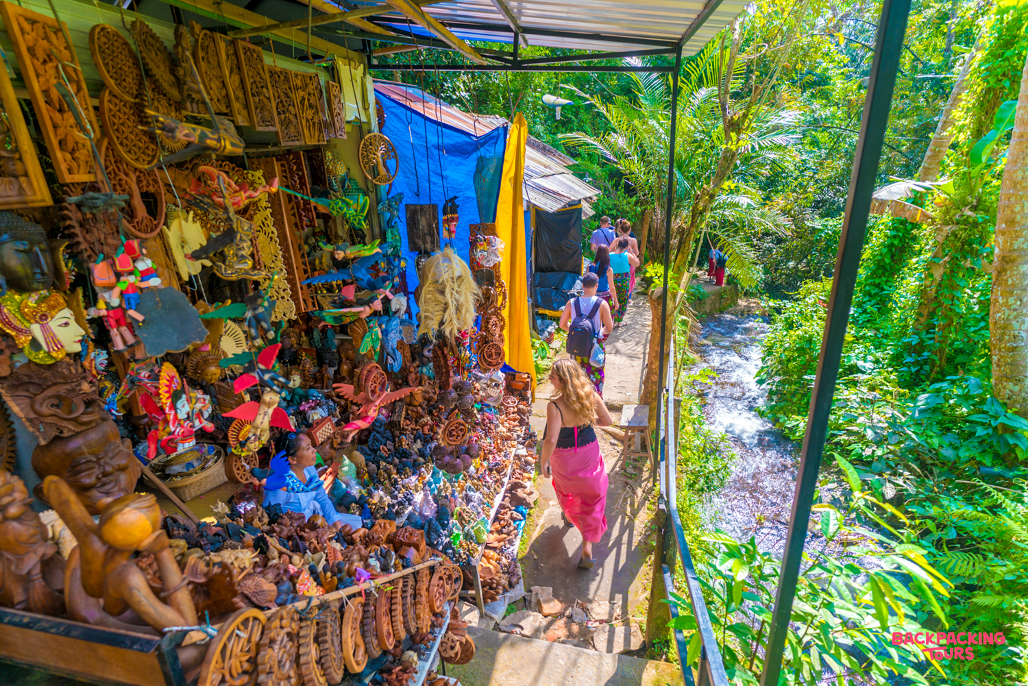Travelers browse a vibrant market stall filled with carved souvenirs along a jungle path in Ubud.
