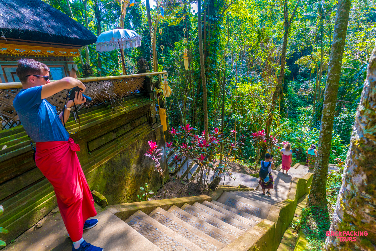 A man in a red sarong photographs a Balinese temple and lush jungle scenery in Ubud, Bali.