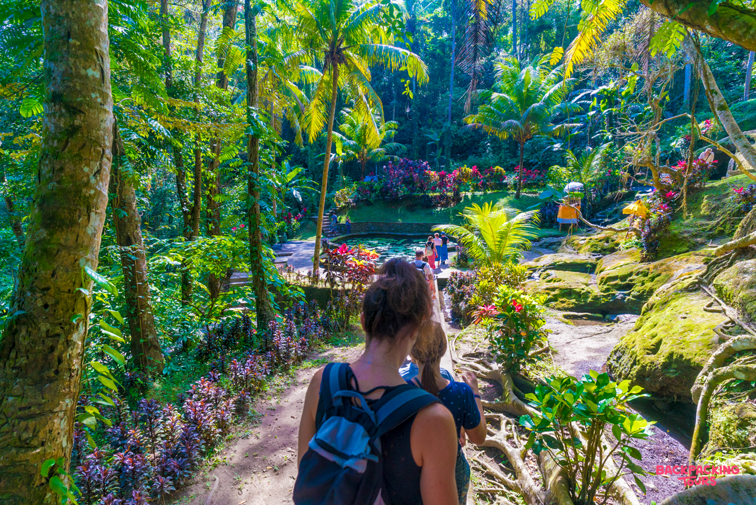 Two travelers explore a vibrant tropical garden path in Ubud, surrounded by lush palms and colorful flowers.