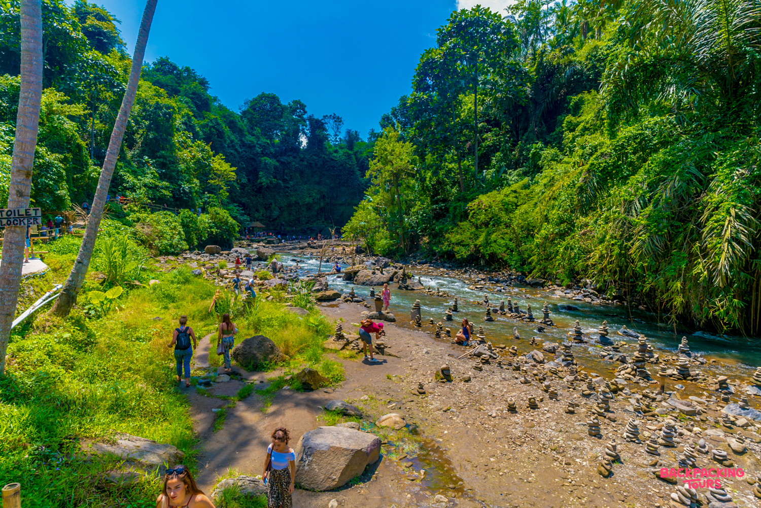 Travelers explore a wide riverbed with rock stacks, surrounded by lush tropical jungle in Bali.