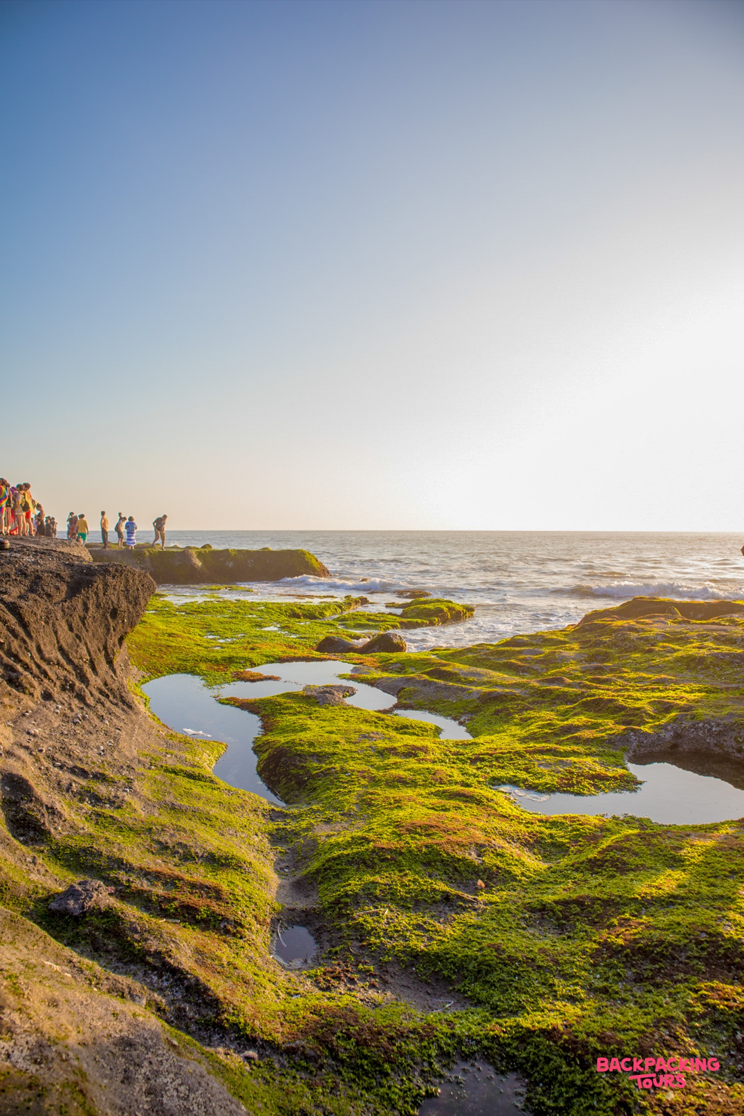 Tourists gather on rocky cliffs covered in green moss, overlooking the ocean at sunset in Bali.
