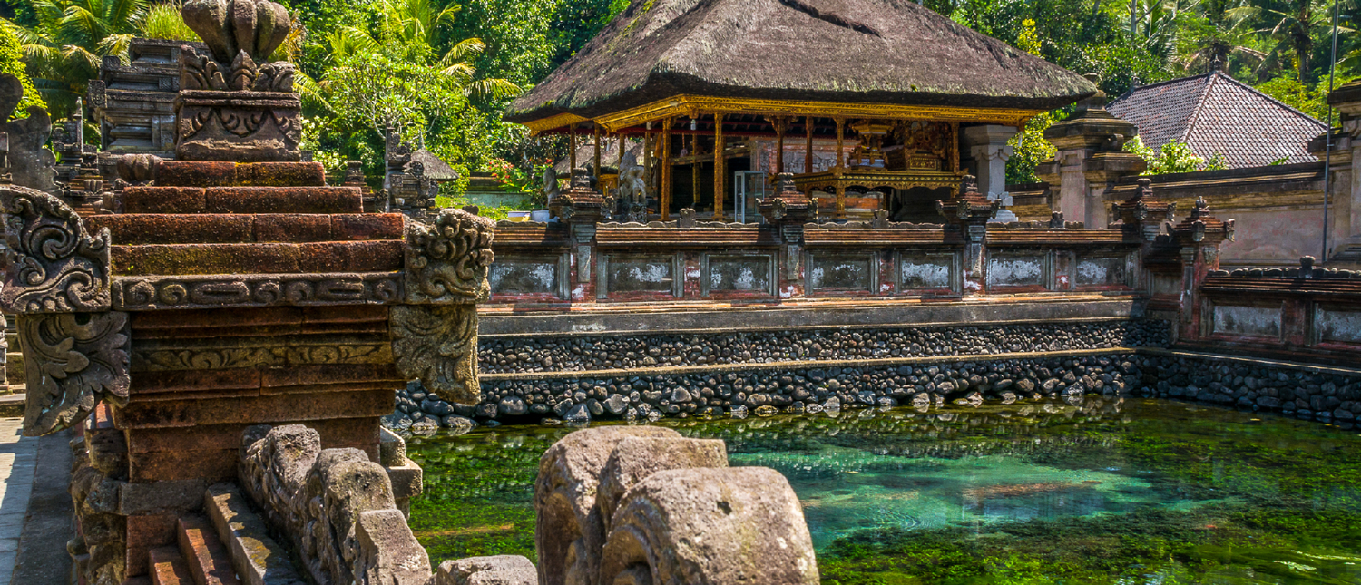 Ornate stone temple structures with thatched roofs beside a clear, green water pond in Ubud, Bali.