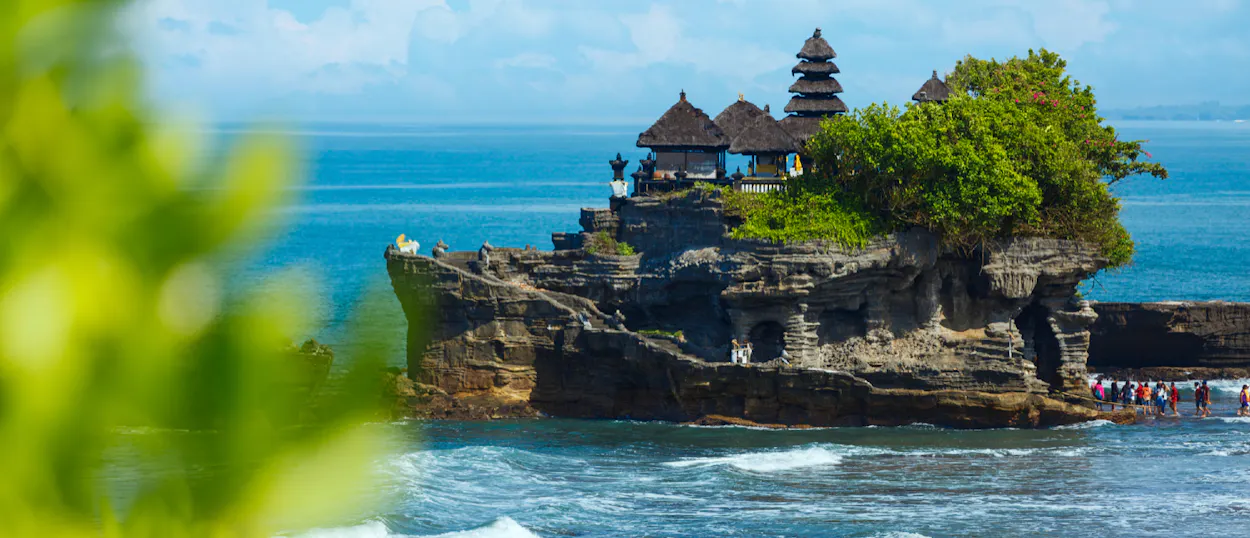 Stunning Balinese temple on a rocky island, surrounded by blue ocean, with people exploring the shore.