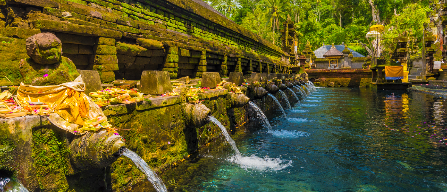 Sacred water flows from moss-covered stone spouts into a clear pool at Tirta Empul temple.