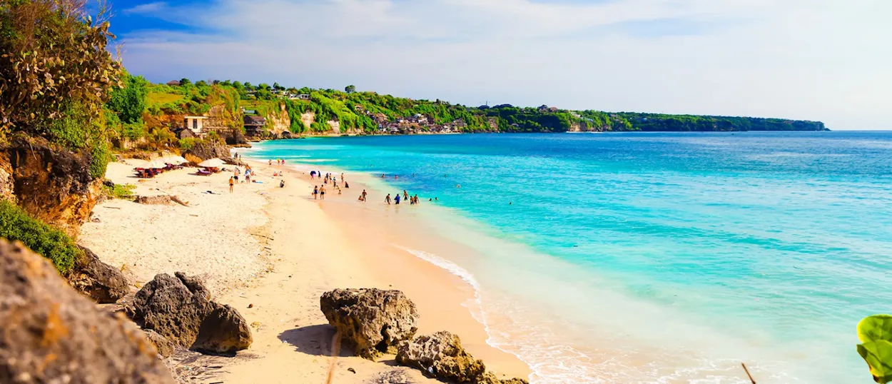 People enjoying the clear turquoise water and sandy beach bordered by green cliffs in Kuta, Bali.