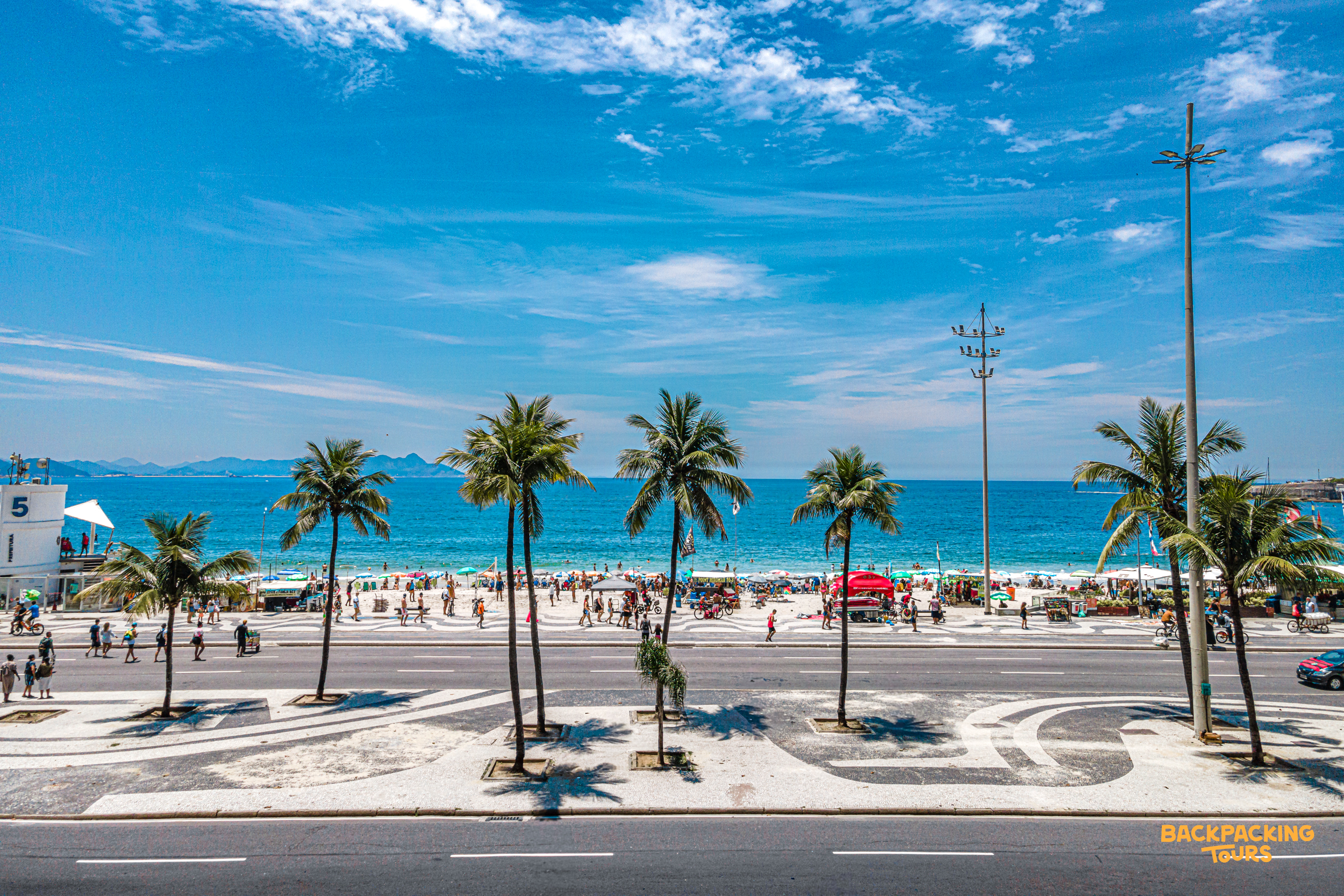 A wide, sunny view of people relaxing on Copacabana Beach, with palm trees and blue ocean.