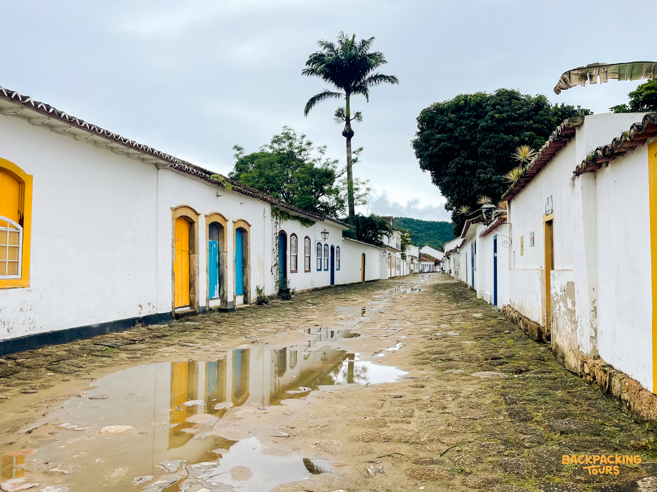 Cobbled street in Paraty with white colonial buildings, colorful doors, and puddles reflecting the sky.