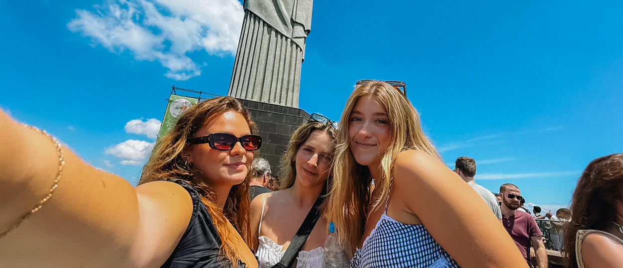 Three smiling women take a selfie in front of the Christ the Redeemer statue in Rio de Janeiro.