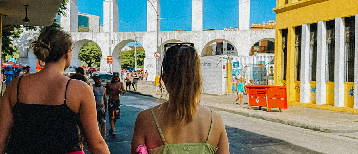 Two women stroll through a lively Rio de Janeiro street, passing the historic Arcos da Lapa.
