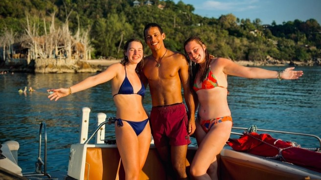 Three smiling young friends in swimsuits on a boat, enjoying a sunny day in a tropical bay.
