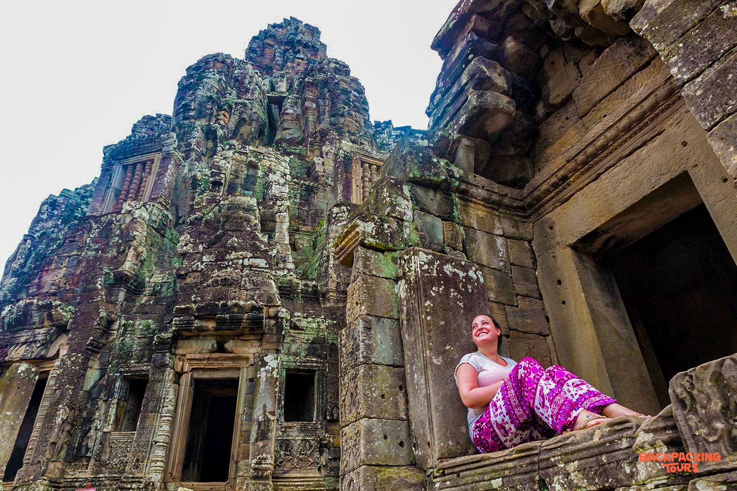 Smiling woman relaxing on a stone ledge, admiring the ancient temple architecture at Angkor Wat.