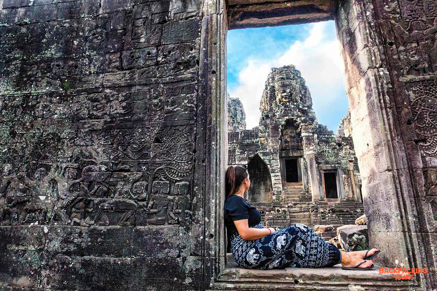 A woman sits in a carved stone doorway, gazing at the ancient Bayon Temple in Siem Reap, Cambodia.
