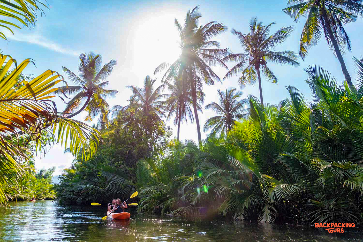 Two people kayak on a serene river surrounded by lush tropical palm trees in Kampot.