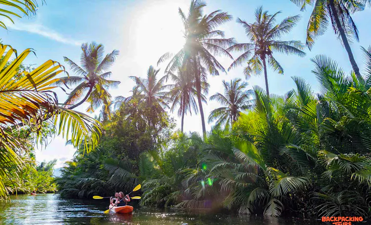 Two people kayak on a calm river, surrounded by lush tropical palm trees under a bright sun in Kampot.