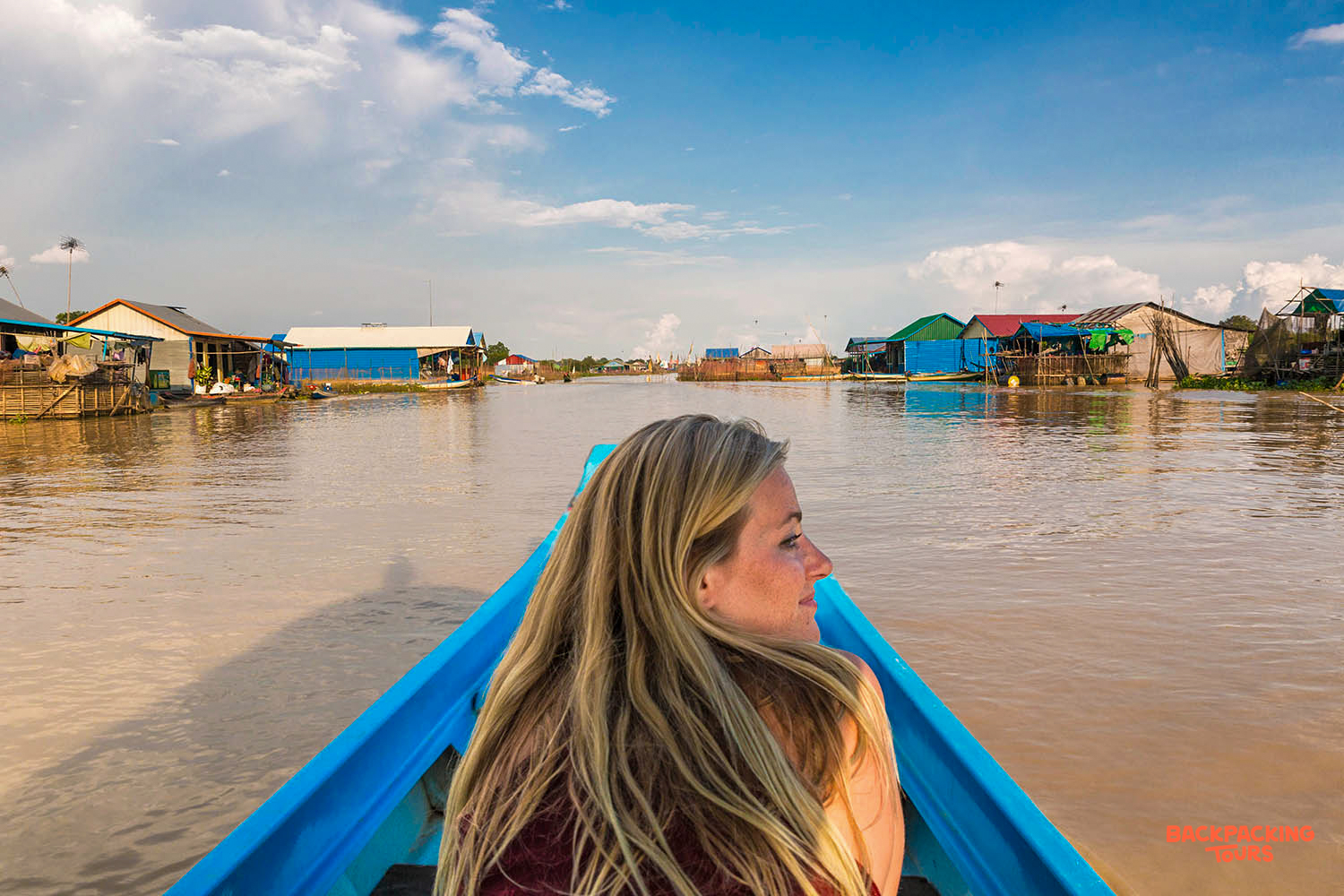 A blonde woman in a blue boat explores traditional floating villages under a blue sky in Siem Reap.