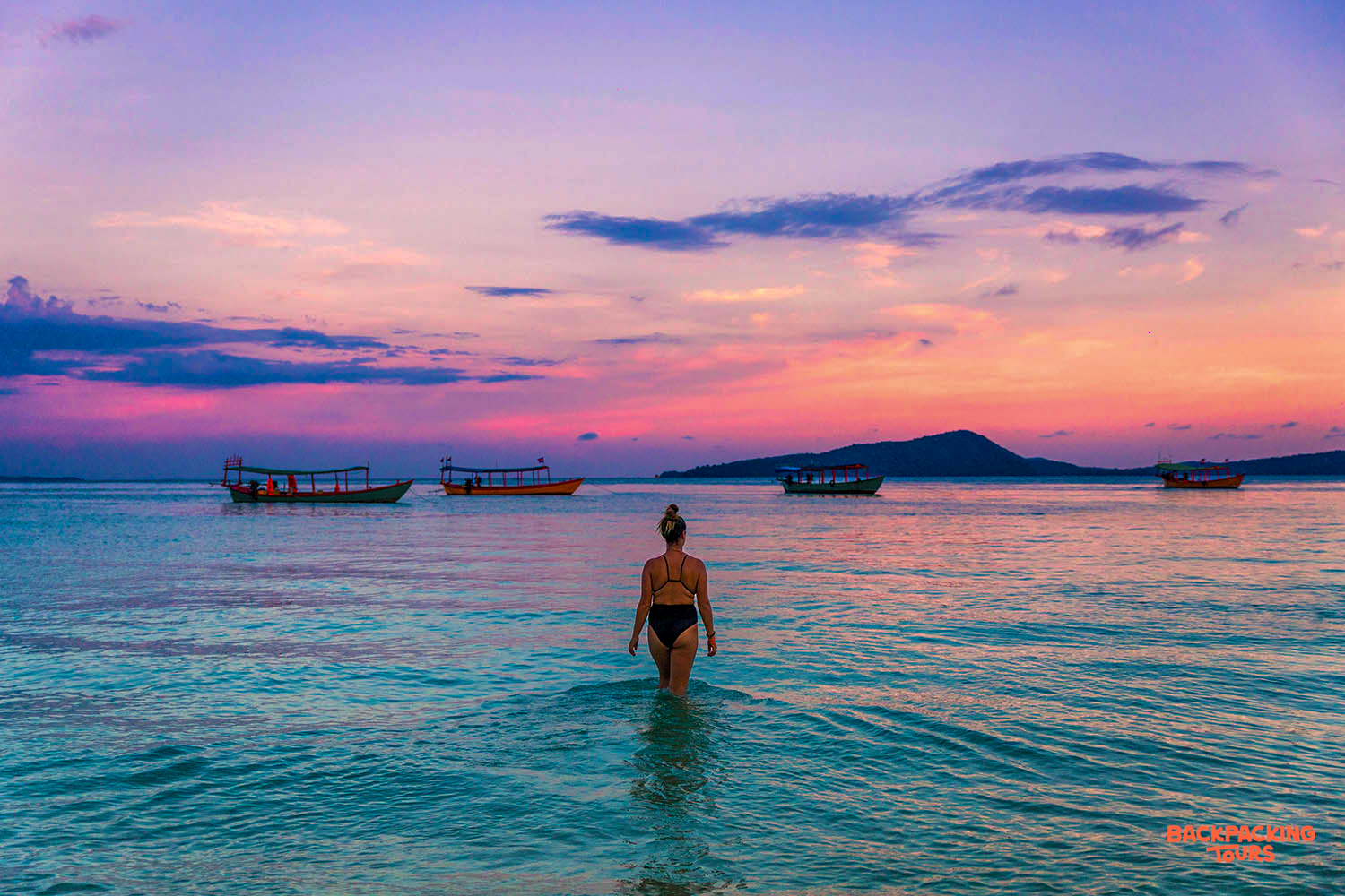 A woman wades into the calm waters of Koh Rong, enjoying a stunning sunset with colorful skies.