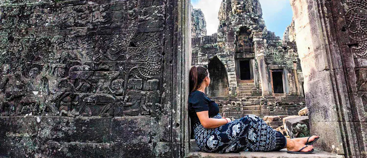 A traveler sits on an intricately carved stone wall, gazing at the majestic Bayon Temple in Cambodia.
