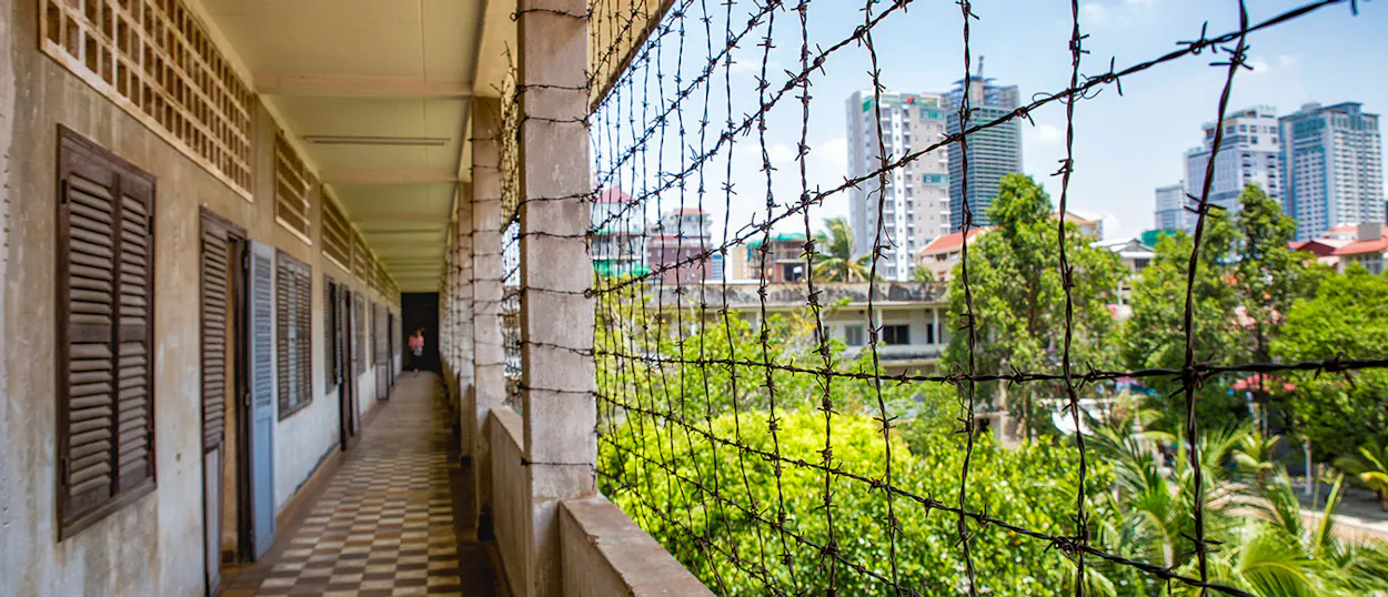 A long, somber corridor with barbed wire fencing, overlooking green trees and Phnom Penh city.