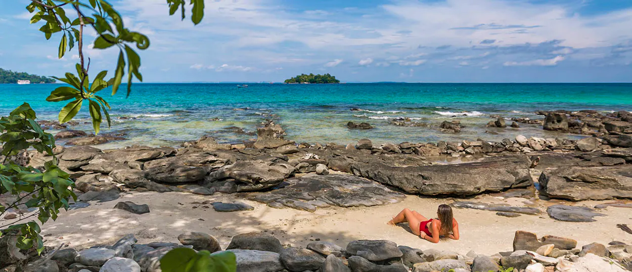 A woman in a red bikini relaxes on a rocky beach by the turquoise ocean in Koh Rong.