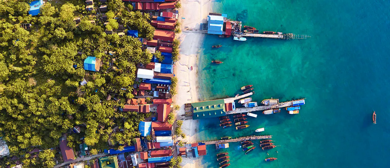 Aerial view of Koh Rong's vibrant coastline, with a sandy beach, lush trees, and longtail boats in turquoise water.