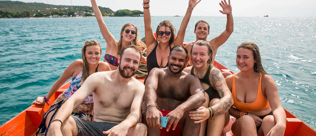A group of happy backpackers waving from a red longtail boat on the clear waters of Koh Rong.